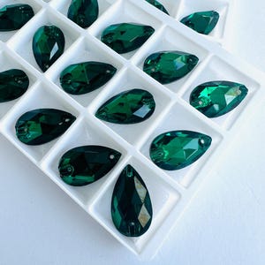 May include: A selection of emerald green, teardrop-shaped crystals displayed in a white tray. The faceted surfaces of the gems catch the light, highlighting their rich green hue. Suitable for jewellery making and various craft applications.