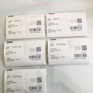 May include: Several small, white packets, each marked with "SWAROVSKI" and product information. The packets contain crystals, with details such as "Rose FB" and "XILION Rose" visible. Each packet has a unique code and a crystal diagram.