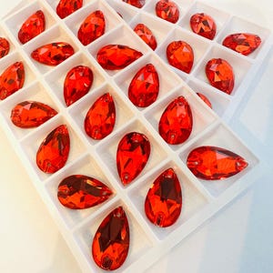 May include: A collection of faceted, red teardrop-shaped rhinestones arranged in a white divided tray. The rhinestones have a glossy finish and are of varying sizes, with a hole at the top for stringing or attachment.