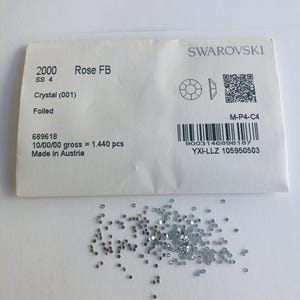 Puede incluir: Un paquete de papel blanco etiquetado como Swarovski 2000 Rose FB SS 4 Crystal (001) Foiled, que contiene 1.440 piezas, con un código de barras y un código QR. Debajo del paquete hay un montón disperso de pequeños cristales redondos facetados.