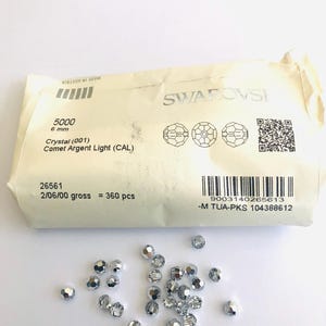 May include: A package of Swarovski crystals, labeled "5000 6 mm Crystal (001) Comet Argent Light (CAL)". A scattering of silver-colored, faceted crystals are visible. The package has a barcode and the text "MADE IN AUSTRIA".