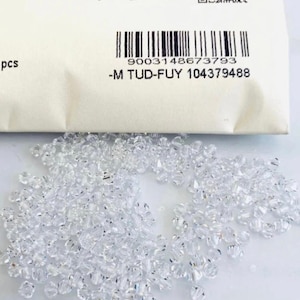 May include: A collection of clear, faceted crystal beads scattered on a white surface. A white packet with a barcode and the text "-M TUD-FUY 104379488" is in the background. The beads are small and reflect light.
