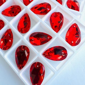 Pode incluir: Uma coleção de strass vermelhos facetados em forma de gota, dispostos em um organizador de plástico branco. Cada gema tem uma extremidade pontiaguda e um furo para enfiar. As gemas são de uma cor vermelha profunda e vibrante.