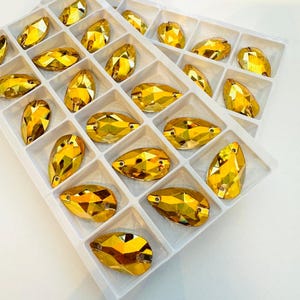 May include: A white tray filled with golden-yellow, faceted rhinestones in a teardrop shape. The rhinestones are arranged in a grid. Each stone has multiple facets, reflecting light. The tray is set against a white background. The rhinestones are suitable for embellishment.