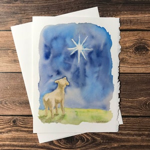May include: A watercolor painting of a white lamb looking up at a bright white star in a blue night sky. The lamb is standing on a green field.