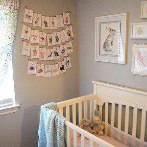 May include: A nursery interior with a white cot, blue blanket, and soft toys. An alphabet banner with animal illustrations is suspended above. A framed watercolour of a rabbit and other artwork adorn the grey walls, creating a charming space.