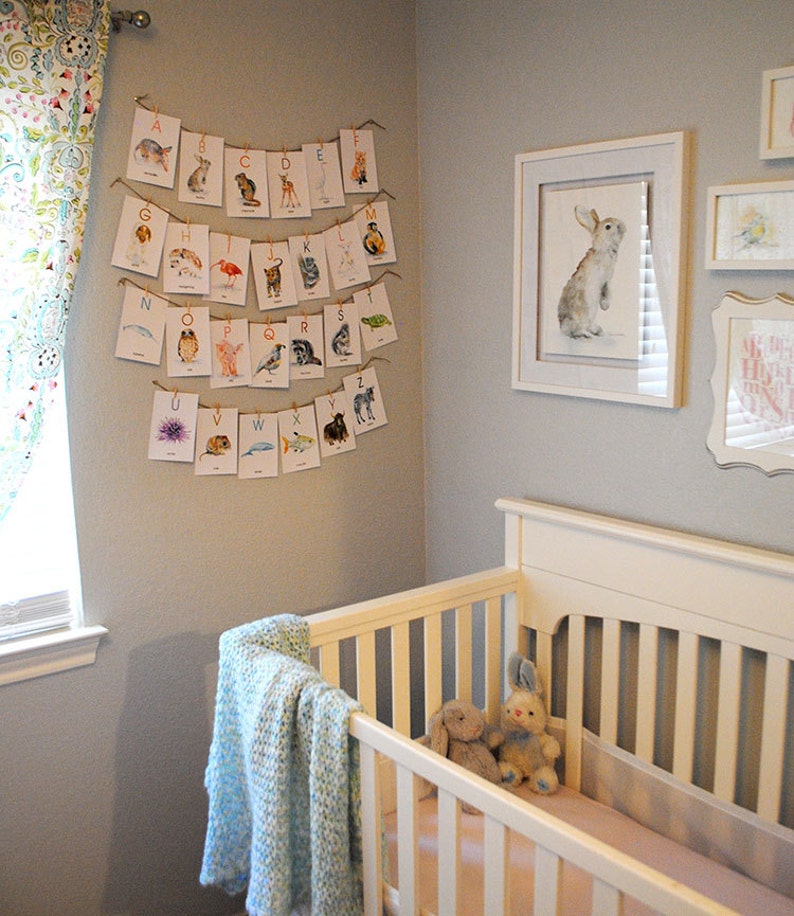 May include: A nursery interior with a white cot, blue blanket, and soft toys. An alphabet banner with animal illustrations is suspended above. A framed watercolour of a rabbit and other artwork adorn the grey walls, creating a charming space.