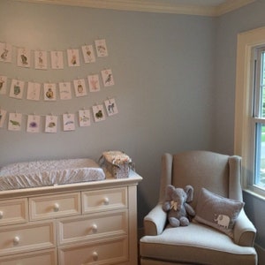 May include: A nursery with a pale blue wall, a white chest of drawers, and a beige armchair. An alphabet banner hangs above the changing table. An elephant soft toy and a cushion with an elephant graphic are on the chair.