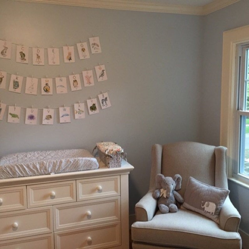 May include: A nursery with a pale blue wall, a white chest of drawers, and a beige armchair. An alphabet banner hangs above the changing table. An elephant soft toy and a cushion with an elephant graphic are on the chair.