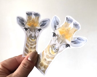 May include: Two watercolor giraffe stickers with a white background. The giraffes are looking directly at the camera with their heads tilted slightly. The stickers are printed on a clear, glossy paper.