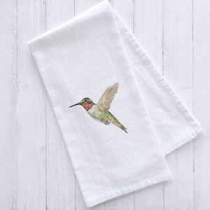 May include: White kitchen towel with a watercolor illustration of a hummingbird in flight. The hummingbird is green, brown, and red with a long beak.