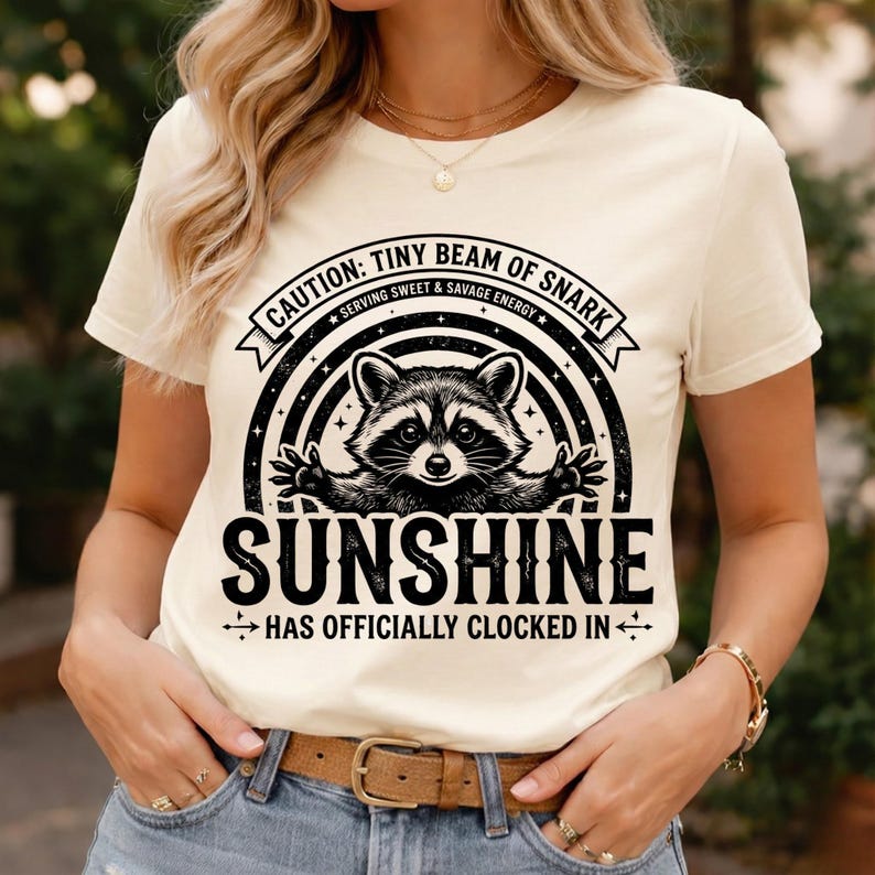 May include: Cream-colored t-shirt featuring a raccoon graphic with the text "CAUTION: TINY BEAM OF SNARK" above and "SUNSHINE HAS OFFICIALLY CLOCKED IN" below. The design includes a black and white raccoon illustration.