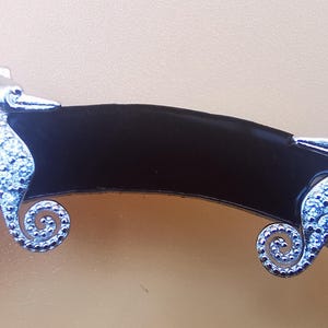 May include: A pair of silver seahorse earrings. Each earring features a seahorse design encrusted with small, clear crystals. The seahorses are positioned on a dark, curved surface against a light brown background.