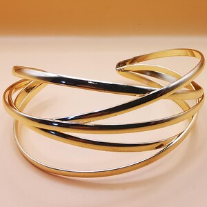 Può includere: Un bracciale a polsino color oro con un design a più strati e a spirale. Il bracciale presenta diverse fasce curve che si sovrappongono e si intrecciano, creando un aspetto aperto ed elegante. Il gioiello è posto su uno sfondo color pesca chiaro.
