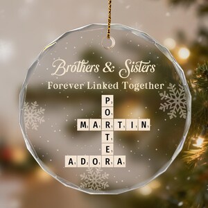 May include: A clear, round Christmas ornament with the words "Brothers & Sisters Forever Linked Together." Scrabble-style tiles spell out names. The ornament hangs from a gold string, with snowflakes and a Christmas tree in the background.