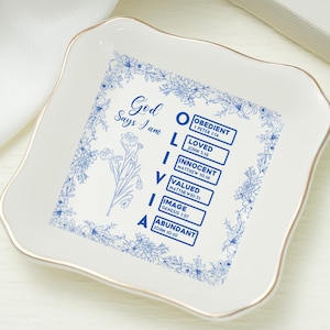 Puede incluir: Una bandeja de cerámica blanca con borde dorado, con diseños florales azules y texto. El plato tiene las palabras "Dios dice que soy" y una lista de afirmaciones positivas. Un anillo de oro con una perla está en la imagen.