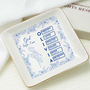 Puede incluir: Una bandeja cuadrada de cerámica blanquecina con borde dorado. La bandeja presenta diseños florales azules y el texto "God Says I am", junto con el nombre "Olivia" y palabras relacionadas. Un anillo está en primer plano.