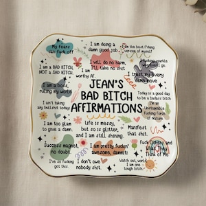 Könnte beinhalten: Eine weiße dekorative Keramikplatte mit goldenem Rand, mit dem Text "Jean's Bad Bitch Affirmations". Die Platte ist mit Affirmationen und farbenfrohen Illustrationen verziert. Die Platte ist ca. 20 cm lang.