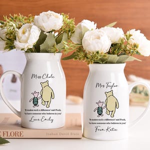May include: Two white ceramic vases with handles, each holding white flowers. The vases feature a Winnie the Pooh design with text that reads "Miss Chole" and "Mrs Taylor". The text also includes a quote and a name.