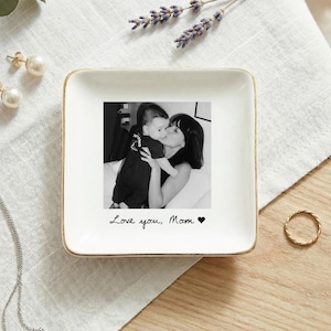 May include: A white square trinket dish with a gold rim features a black and white photo of a mother and child. The dish has the handwritten text "Love you, Mom" and a heart symbol. A gold ring and pearl earrings are nearby.