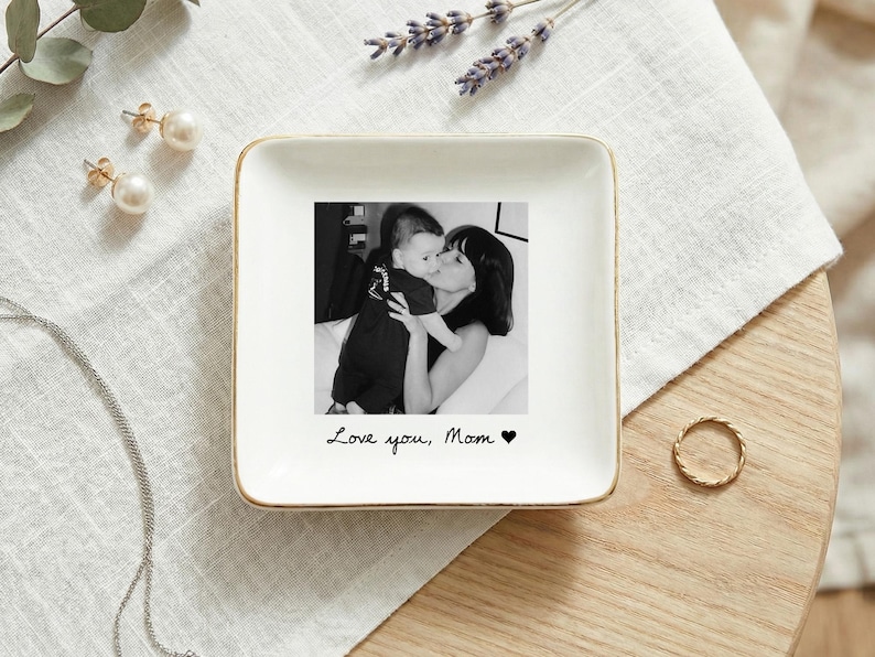 May include: A white square trinket dish with a gold rim features a black and white photo of a mother and child. The dish has the handwritten text "Love you, Mom" and a heart symbol. A gold ring and pearl earrings are nearby.