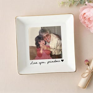 May include: A square, white ceramic trinket dish with a gold rim. The center features a photograph of a woman and a child. The words "Love you, grandma" are written below the photo. A pink flower and a rolled scroll are in the background.