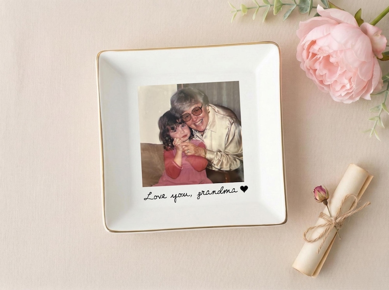 May include: A square, white ceramic trinket dish with a gold rim. The center features a photograph of a woman and a child. The words "Love you, grandma" are written below the photo. A pink flower and a rolled scroll are in the background.
