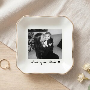May include: A white ceramic trinket dish with a gold rim, featuring a black and white photo of a mother and baby. The text "Love you, Mom" is written below the photo, with a heart symbol. A gold ring and flowers are also visible.