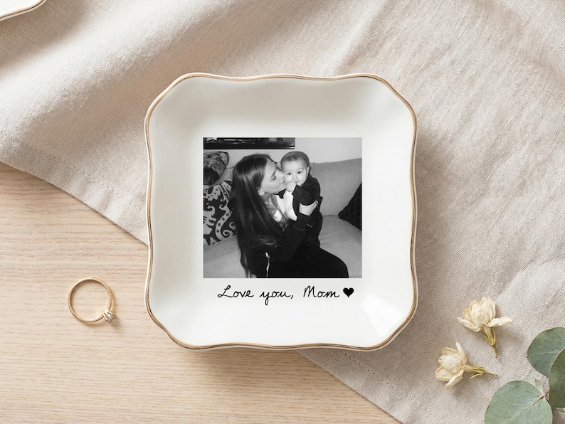 May include: A white ceramic trinket dish with a gold rim, featuring a black and white photo of a mother and baby. The text "Love you, Mom" is written below the photo, with a heart symbol. A gold ring and flowers are also visible.