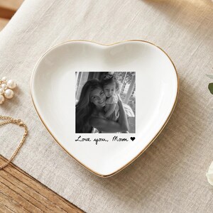 May include: A heart-shaped white ceramic trinket dish with a gold rim. The dish features a black and white photo and the text "Love you, Mom" with a heart symbol. A gold bracelet and pearl earrings are next to the dish.