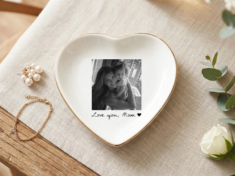 May include: A heart-shaped white ceramic trinket dish with a gold rim. The dish features a black and white photo and the text "Love you, Mom" with a heart symbol. A gold bracelet and pearl earrings are next to the dish.