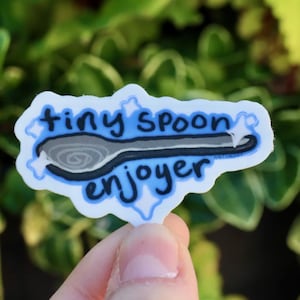 Pode incluir: Um autocolante com as palavras "tiny spoon enjoyer" em azul e preto, com um desenho de uma colher. O autocolante é apresentado sobre um fundo verde desfocado.