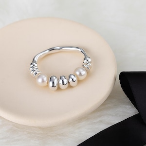 Può includere: Un anello d'argento dal design unico. L'anello presenta una serie di piccole perle rotonde e perline d'argento allungate. L'anello è esposto su una superficie color crema con un nastro nero sullo sfondo.