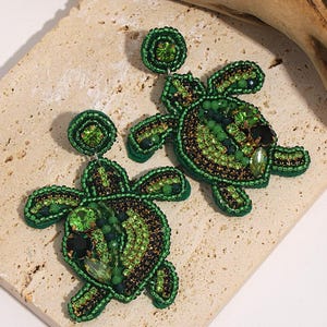 May include: A pair of green and black beaded turtle earrings. The earrings have a detailed design with various shades of green beads and accents, creating a textured appearance. The earrings are set against a neutral background.