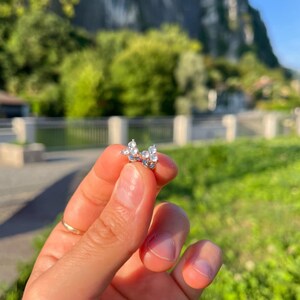 Può includere: Un piccolo orecchino a lobo argentato con un design a farfalla, ornato da pietre preziose chiare e scintillanti. Lo sfondo è una scena sfocata con alberi verdi e una montagna.