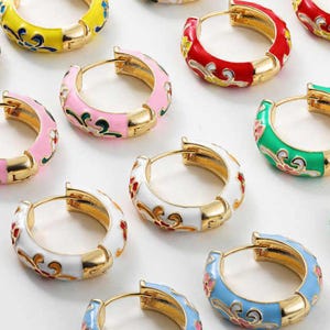 Può includere: Una collezione di orecchini a cerchio smaltati colorati. Gli orecchini presentano metallo color oro con sezioni di smalto rosa, rosso, bianco, verde, giallo e blu. Ogni orecchino ha un design floreale.