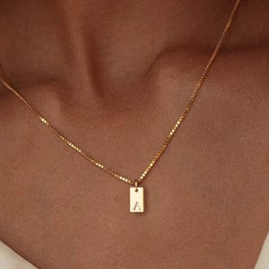 Può includere: Una collana color oro con un ciondolo rettangolare. Il ciondolo ha incisa la lettera "A". La collana ha una catena sottile e il ciondolo è sospeso al centro.
