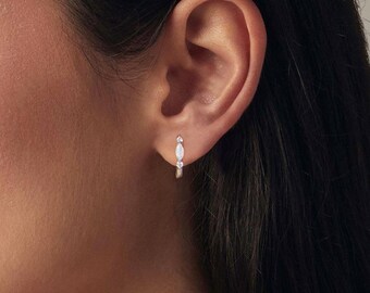 Puede incluir: Primer plano de un pendiente de aro plateado con una ópalo blanco ovalado y una pequeña piedra transparente. Se muestra en una oreja, con la oreja y el pelo oscuro visibles en el fondo.