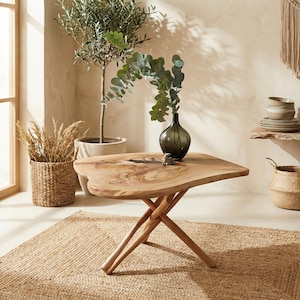 May include: A wooden, free-form coffee table with a natural wood grain finish. The table has a unique, organic shape and is supported by a crossed-leg base. A dark green vase with greenery sits on top. The table is set on a woven jute rug.