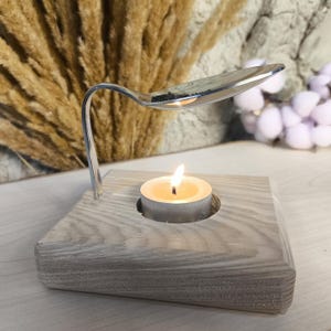 Può includere: Un portacandele in legno con una piccola candela a té che brucia all'interno. Un cucchiaio di metallo piegato è posizionato sopra la candela, creando un design unico e rustico.