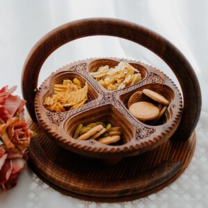 Puede incluir: Una bandeja de madera para servir con cuatro compartimentos en forma de corazón, llenos de aperitivos. La bandeja tiene un diseño floral tallado y un asa. Los aperitivos incluyen patatas fritas, galletas y fruta seca. La bandeja es marrón.