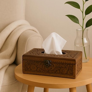 以下が含まれることがあります： 精巧な花柄の彫刻と金属製の留め金が付いた、長方形の濃い茶色の木製ティッシュボックス。白いティッシュが見えます。小さな丸い薄茶色の木製テーブルの上にあり、緑の葉が入ったガラスの花瓶の隣にあります。