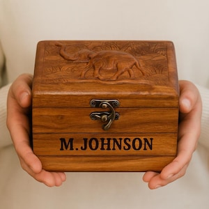 May include: A wooden box with an elephant carving on the lid and the name "M.JOHNSON" engraved on the front. The box has a metal clasp and is made of a warm, brown wood. The elephant carving is detailed and adds a decorative element.