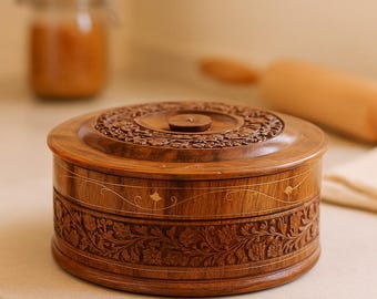 Puede incluir: Una caja redonda de madera con intrincadas tallas florales. La tapa presenta un diseño circular detallado, y la caja tiene una banda decorativa con detalles dorados. La caja es de madera marrón cálida y está sobre una superficie blanca.