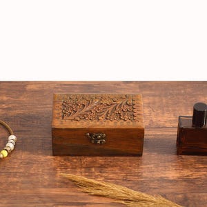 Puede incluir: Una caja de madera con un diseño floral tallado en la parte superior. La caja tiene un cierre de latón y está sentada sobre una superficie de madera. También hay una pequeña botella de perfume y un collar en la superficie.