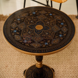 Pu&ograve; includere: Tavolino rotondo con una superficie scura ornata da un motivo floreale dettagliato in oro e blu. Il tavolo ha un bordo e una base dorati e una gamba a colonna. Il tavolo ha un diametro di circa 61 cm.