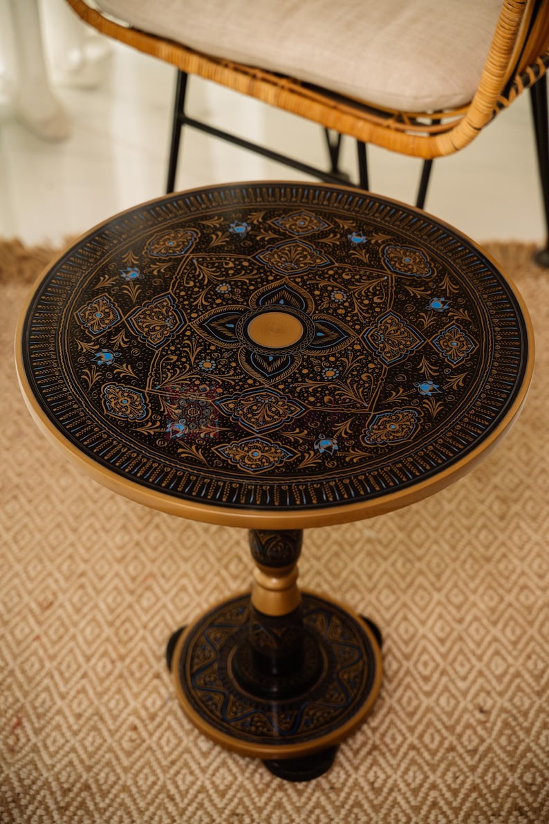 Pu&ograve; includere: Tavolino rotondo con una superficie scura ornata da un motivo floreale dettagliato in oro e blu. Il tavolo ha un bordo e una base dorati e una gamba a colonna. Il tavolo ha un diametro di circa 61 cm.