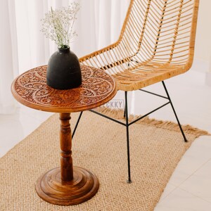 May include: A round, wooden side table with intricate floral carvings. A black vase holds white flowers on the table. A woven chair with black metal legs is behind the table. The table sits on a woven rug.