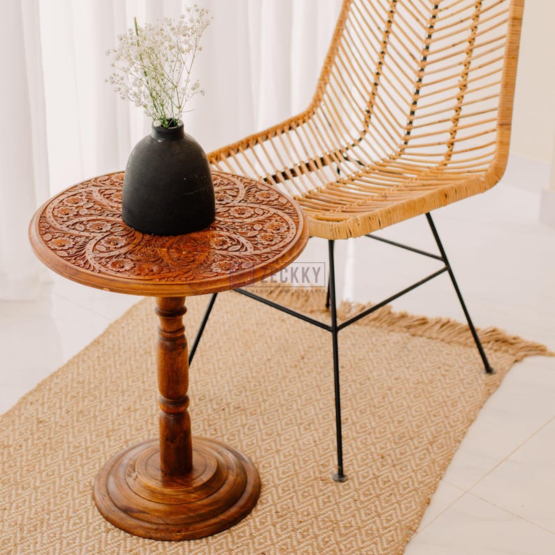 May include: A round, wooden side table with intricate floral carvings. A black vase holds white flowers on the table. A woven chair with black metal legs is behind the table. The table sits on a woven rug.