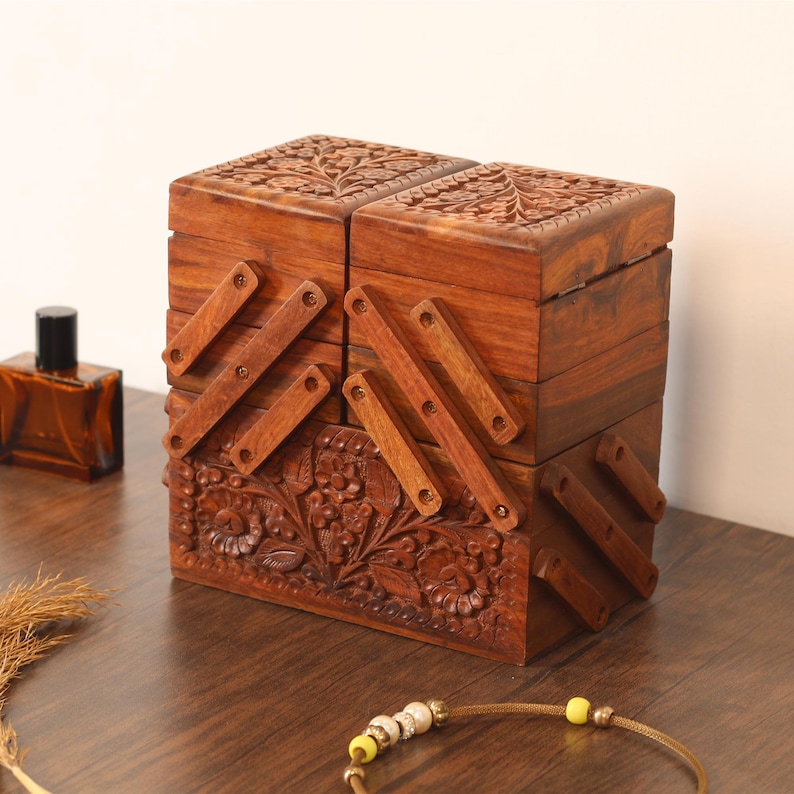 May include: A handcrafted wooden jewelry box with intricate floral carvings. The box is a warm brown color and features multiple compartments that open with wooden supports. A perfume bottle and a necklace are also visible.